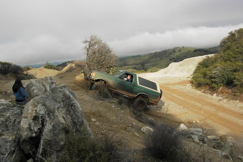 Green SUV with white top on a dirt hill, driver visible, two people watching