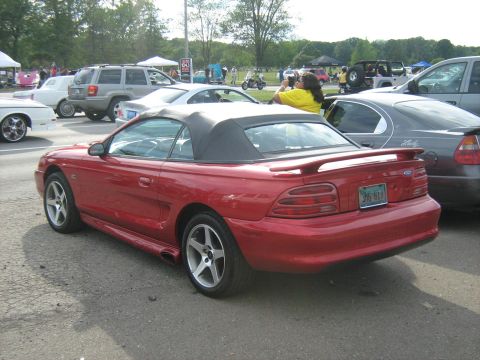 1988 Mercury Grand Marquis - A red convertible sports car is parked in a lot with other vehicles and tents in the background.