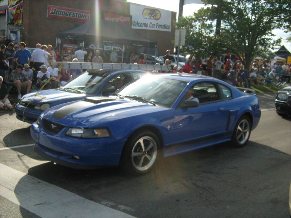Blue Ford Mustang with black stripe on hood, parked next to another car. Crowd watches in background