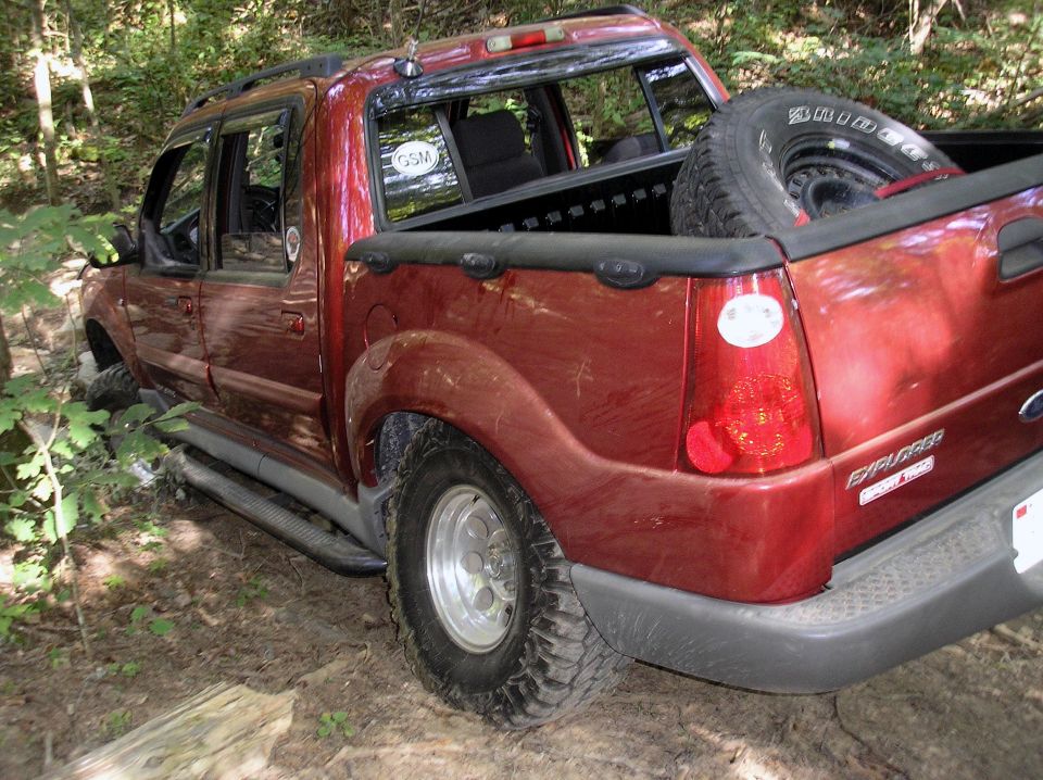 Red pickup truck with a spare tire in the bed, stuck in a wooded area