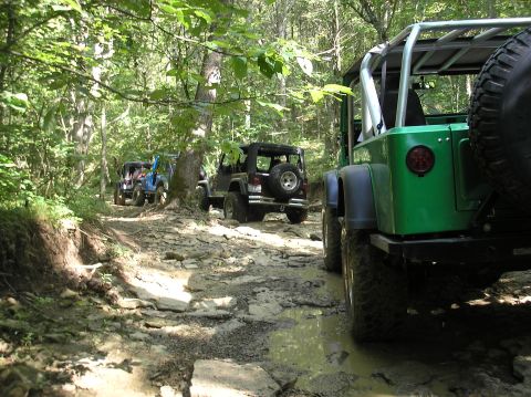 2002 Ford Sport Trac - Several Jeeps navigate a challenging, muddy, and rocky offroad trail through a wooded area.