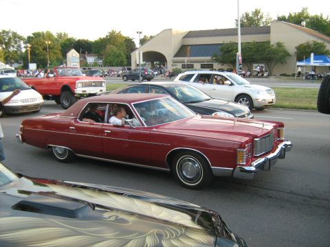 1988 Mercury Grand Marquis - A red sedan cruises down the road, part of a larger car gathering.