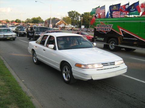 1988 Mercury Grand Marquis - A white sedan cruises down the road, part of a parade or event with other vehicles and spectators.