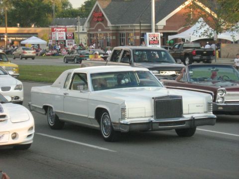 1988 Mercury Grand Marquis - A white Lincoln Continental Mark IV is seen in a parade or car show with other vehicles and spectators.