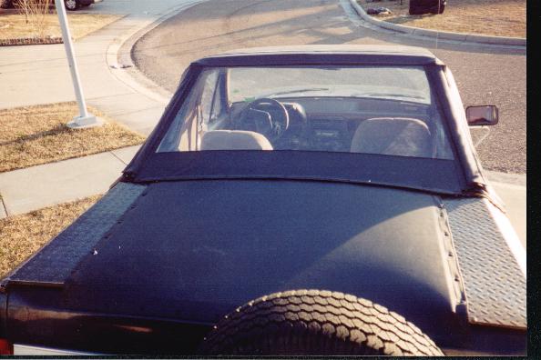 Dark convertible with black soft top and rear-mounted spare tire, parked in a driveway.