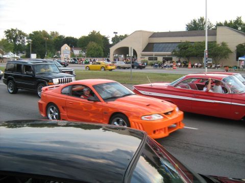1988 Mercury Grand Marquis - An orange Ford Mustang with flame decals is seen in a parade, alongside other vehicles.