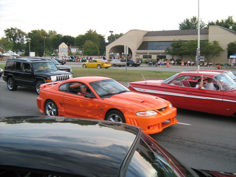 Orange Ford Mustang with flame decals driving in a parade, next to a red classic car