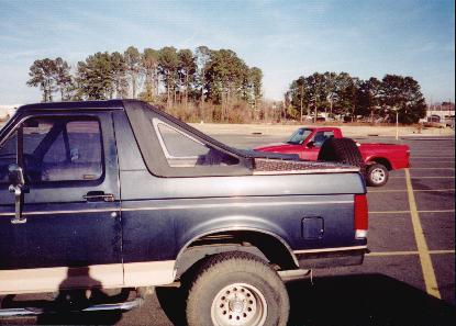 1992 Ford Bronco - A dark blue Ford Bronco is parked in a lot next to a red pickup truck under a clear sky.