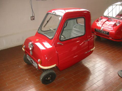 1985 Ford Bronco - A small red microcar is parked indoors next to another vehicle. The license plate is visible.