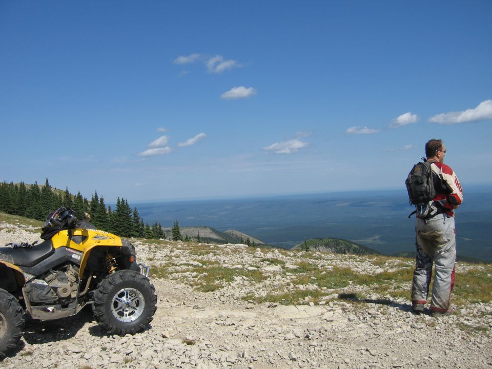 Yellow ATV parked on a rocky mountain trail, man in riding gear standing nearby looking out at the landscape