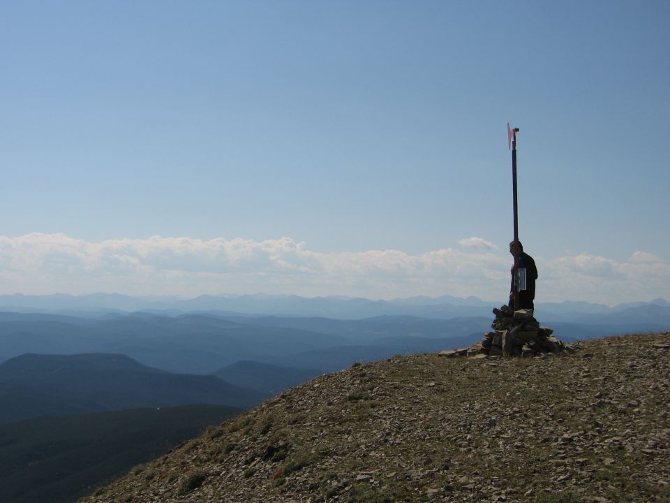 Person on rocky mountain summit near a pole with a flag, with layers of blue mountains in the background under a clear sky