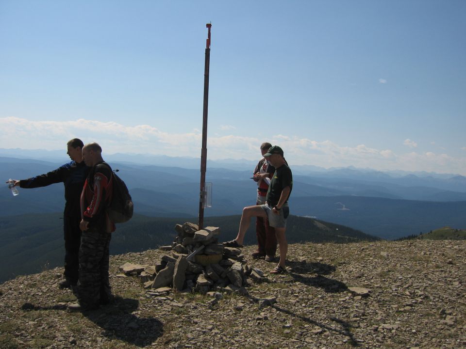 Four people stand on a rocky mountain summit near a pole and cairn, with distant mountains under a blue sky