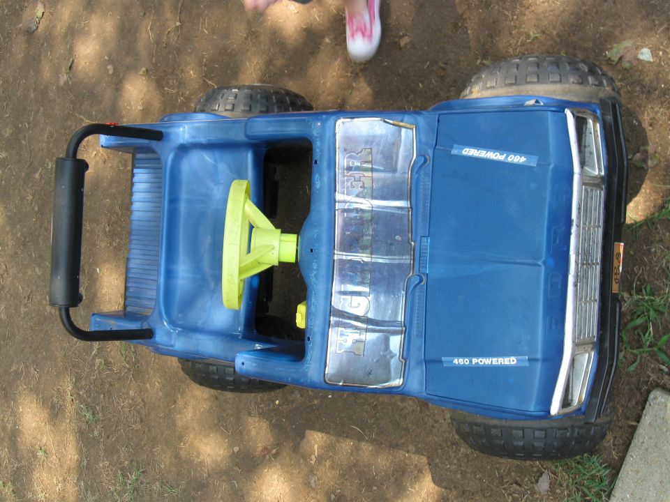Blue toy truck with yellow steering wheel and "460 POWERED" text. A child's legs are visible in the background