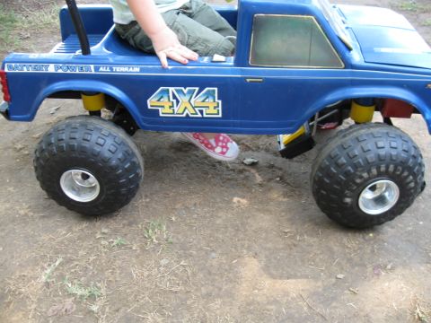 1994 Ford Bronco - A child sits in a blue toy truck with large off-road tires and "4X4" graphics.
