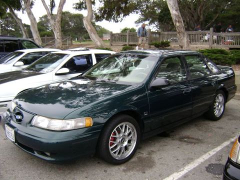 1991 Ford Taurus - A dark green sedan is parked in a lot with other vehicles and trees in the background.