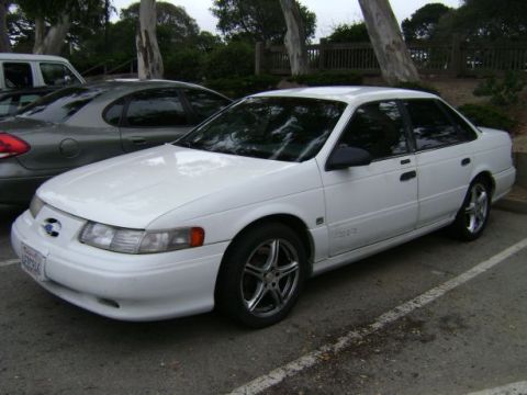 1991 Ford Taurus - A white sedan is parked in a lot next to another car, with trees and a fence in the background.