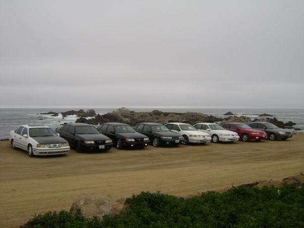 Eight sedans parked on a dirt lot by the ocean.