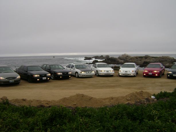 Several sedans parked in a row on a dirt lot by the ocean.
