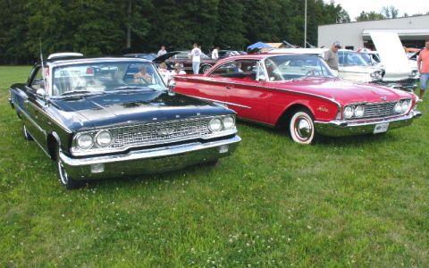 2003 Ford Crown Victoria - Two vintage cars, a black one and a red one, are parked on a grassy field with trees in the background.
