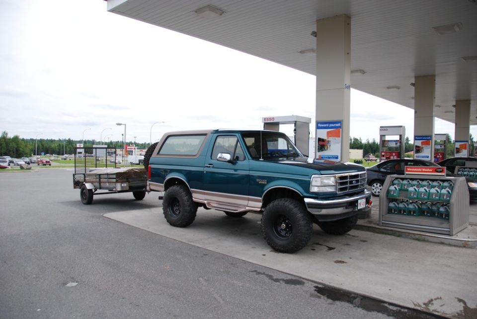 Teal 1996 Ford Bronco with large tires and a trailer parked at a gas station