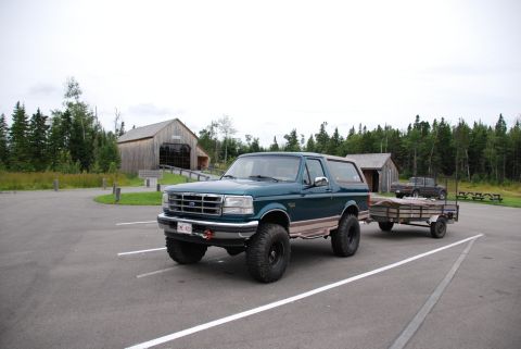 1996 Ford Bronco - A 1996 Ford Bronco is parked in a lot, hitched to a trailer, with wooden buildings and trees in the background.