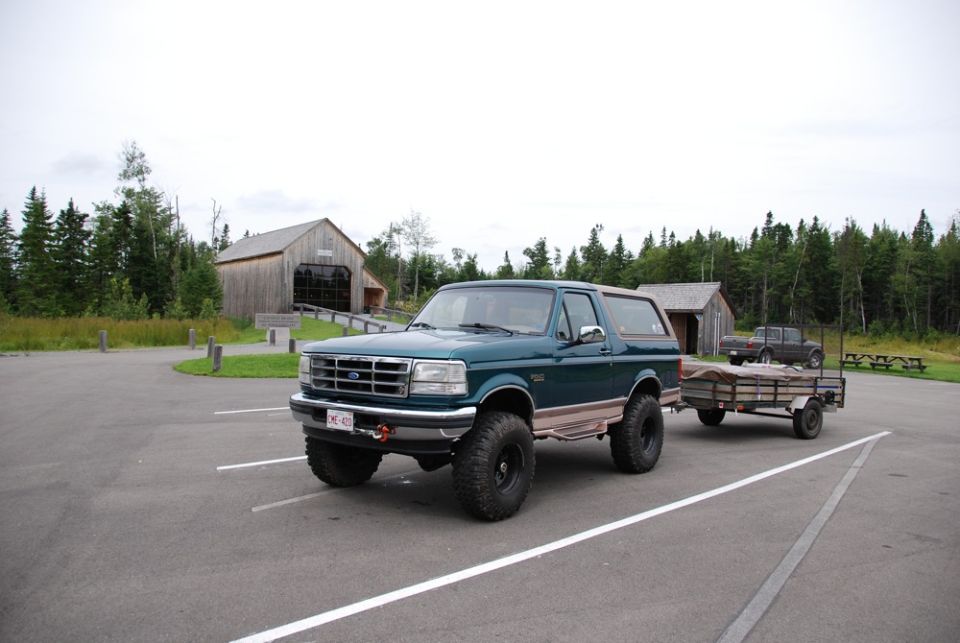 Green 1996 Ford Bronco with lifted suspension and large tires, towing a wooden trailer. License plate visible