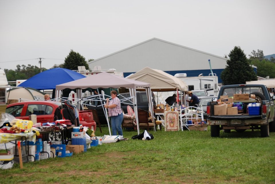 Outdoor market scene with tents, tables of goods, a red car, and a pickup truck loaded with boxes