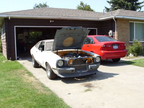 1978 Ford Mustang II - A white coupe with its hood open sits in a driveway next to a red sedan.