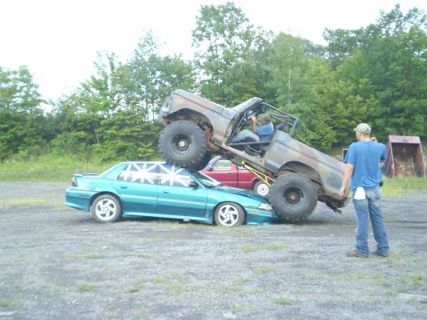 1979 Ford Bronco - A monster truck is shown crushing a teal sedan, while another vehicle and a person watching are also visible in the scene.