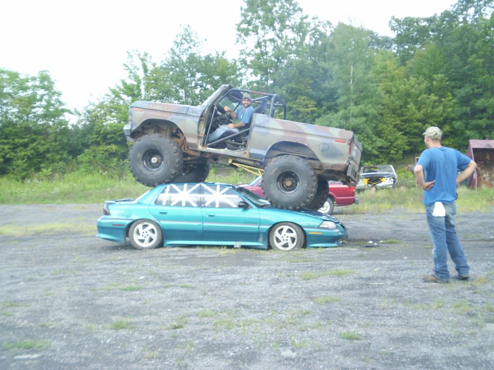 1979 Ford Bronco on top of a teal sedan, with a man standing nearby
