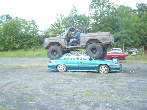 1979 Ford Bronco - A lifted truck is crushing a sedan in an outdoor lot with trees in the background.