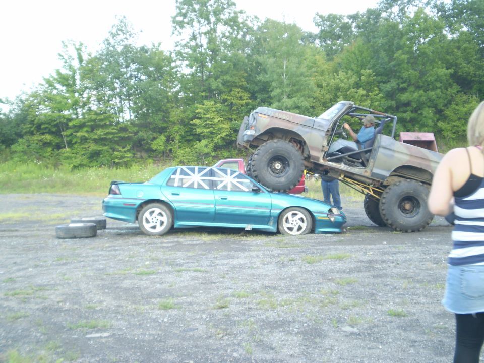 Lifted off-road truck crushing a teal sedan. A person is in the driver's seat of the truck