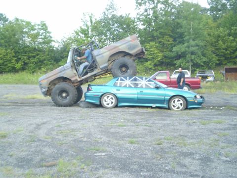 1979 Ford Bronco - A lifted truck is crushing a sedan in an outdoor lot with trees in the background.