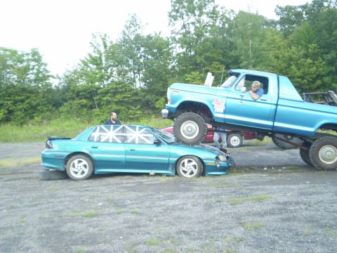 1979 Ford Bronco - A lifted blue pickup truck is crushing a teal sedan that has Union Jack patterns on its windows.