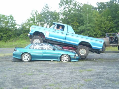 1979 Ford Bronco - A blue pickup truck is shown crushing a teal sedan, with another vehicle in the background.