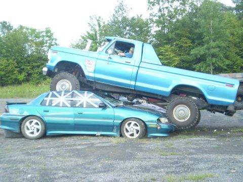 1979 Ford Bronco - A blue pickup truck is crushing a teal sedan, with trees in the background and a watermark at the bottom.