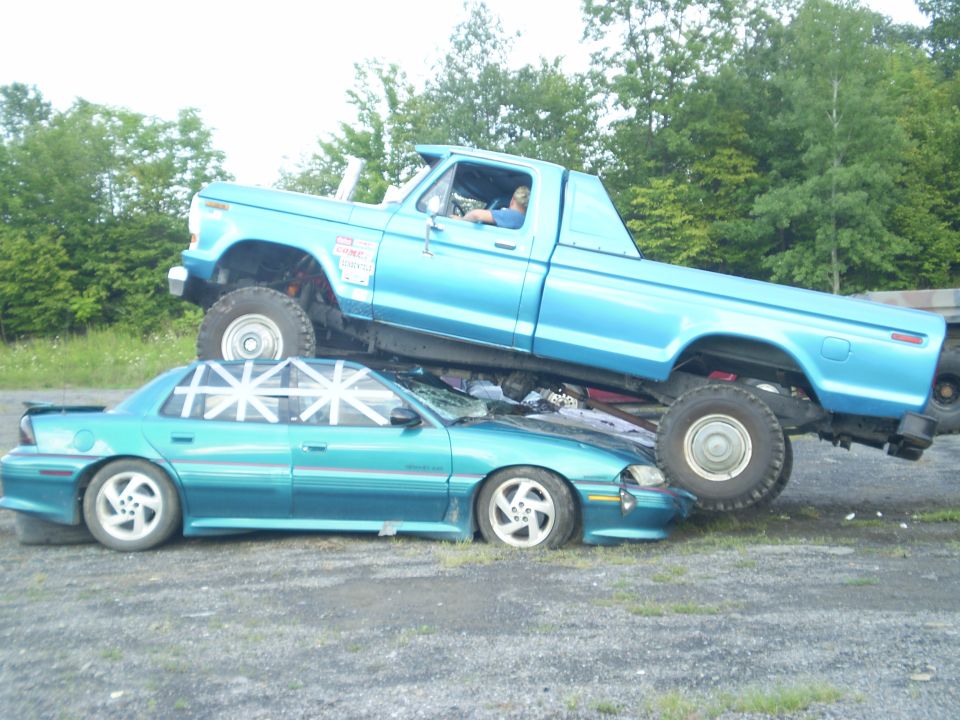 Blue pickup truck is on top of a teal sedan. The truck has a sticker on the door
