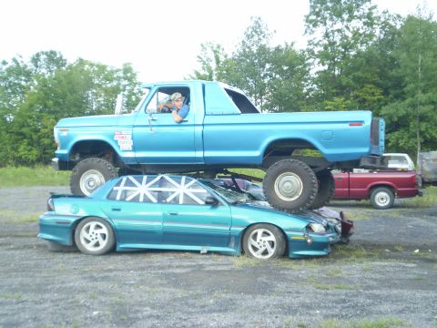 1979 Ford Bronco - A blue 1979 Ford Bronco is shown crushing a teal sedan, with another red truck visible in the background.