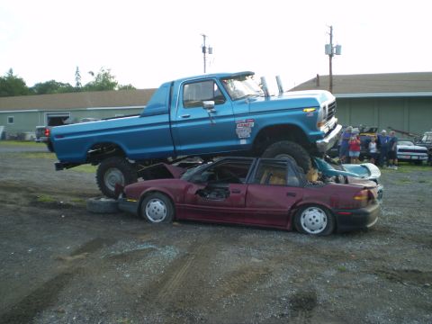 1979 Ford Bronco - A lifted blue pickup truck is parked on top of a wrecked red convertible, with other vehicles and people in the background.