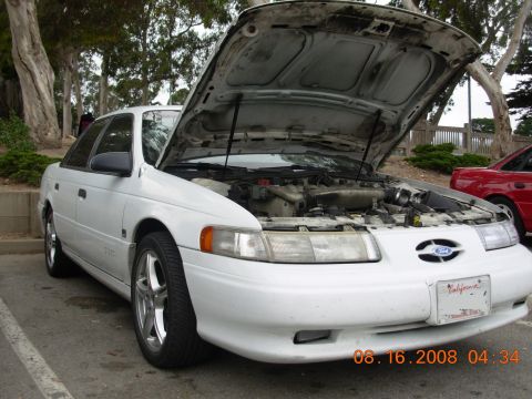 1995 Ford Taurus - A white 1995 Ford Taurus is parked with its hood open, revealing the engine compartment. A red car is parked nearby.