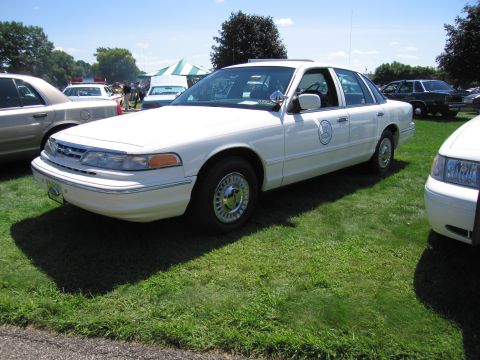 1991 Ford LTD Crown Victoria - white Ford Crown Victoria sedan with a circular door seal and spoke wheels parked on grass among other cruisers.