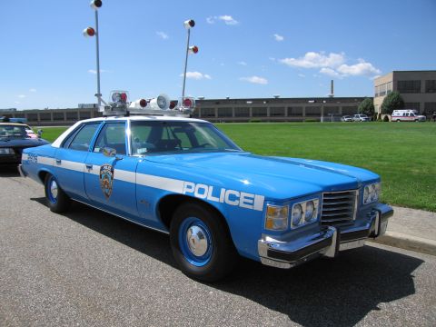 1991 Ford LTD Crown Victoria - Front three-quarter view of a blue and white 1970s Pontiac POLICE sedan with roof beacons and quad headlights.