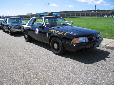 1991 Ford LTD Crown Victoria - black Fox-body Ford Mustang coupe lettered STATE TROOPER with a roof lightbar and spotlight parked on pavement.