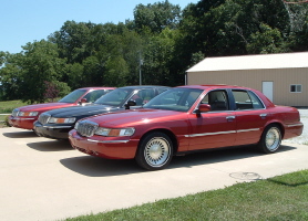 2001 Mercury Grand Marquis - Three Mercury Grand Marquis sedans are lined up in front of a building on a sunny day.