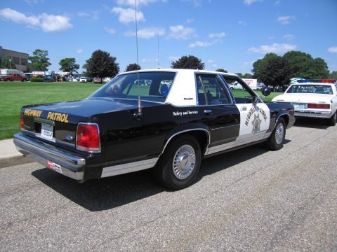 1991 Ford LTD Crown Victoria - Rear three-quarter view of a black-and-white Highway Patrol Ford sedan marked 'Safety and Service' with a gold star on the rear door.