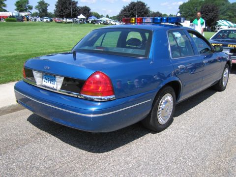 1991 Ford LTD Crown Victoria - Rear three-quarter view of a blue Ford Crown Victoria police sedan with roof light bar parked on pavement near a grassy field.