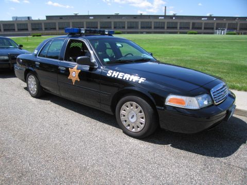 1991 Ford LTD Crown Victoria - black Ford Crown Victoria Sheriff sedan with a gold star door emblem and roof light bar parked beside a green field.