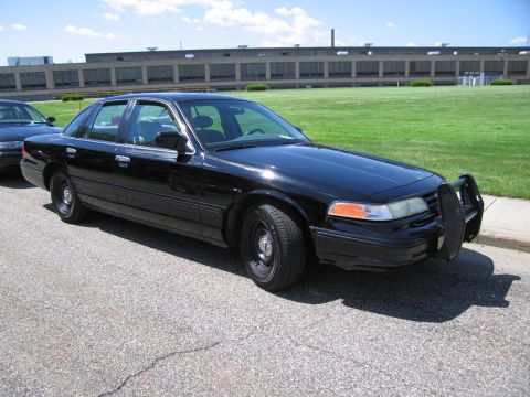 1991 Ford LTD Crown Victoria - unmarked all-black Ford Crown Victoria sedan with a front push bar and black steel wheels parked by a grassy lot.