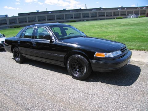 1991 Ford LTD Crown Victoria - unmarked all-black Ford Crown Victoria sedan with black steel wheels parked on pavement in front of a green field.