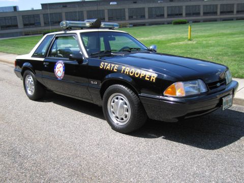 1991 Ford LTD Crown Victoria - black Ford Mustang 5.0 coupe marked 'State Trooper' with a roof light bar parked beside a grassy field.